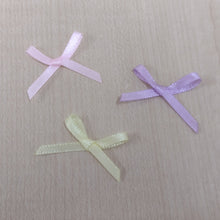 Three small ribbon bows in light pink, lavender, and pale yellow on a light wood surface.