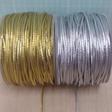 Two spools of thin metallic cord, one gold and one silver, placed on a light wood surface with a green cutting mat in the background.